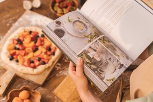 A person reads a cookbook while preparing a fruit tart on a wooden table.